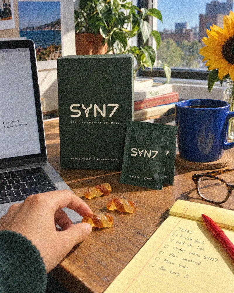 A hand picks up a twist gummy from a sunny desk with a laptop, notepad, blue mug, sunflowers and the SYN7 box