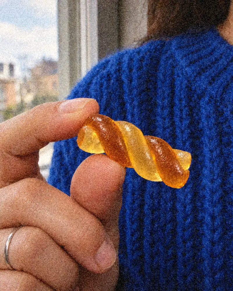 Fingers hold one amber and yuzu twist gummy in front of a cobalt blue knit sweater