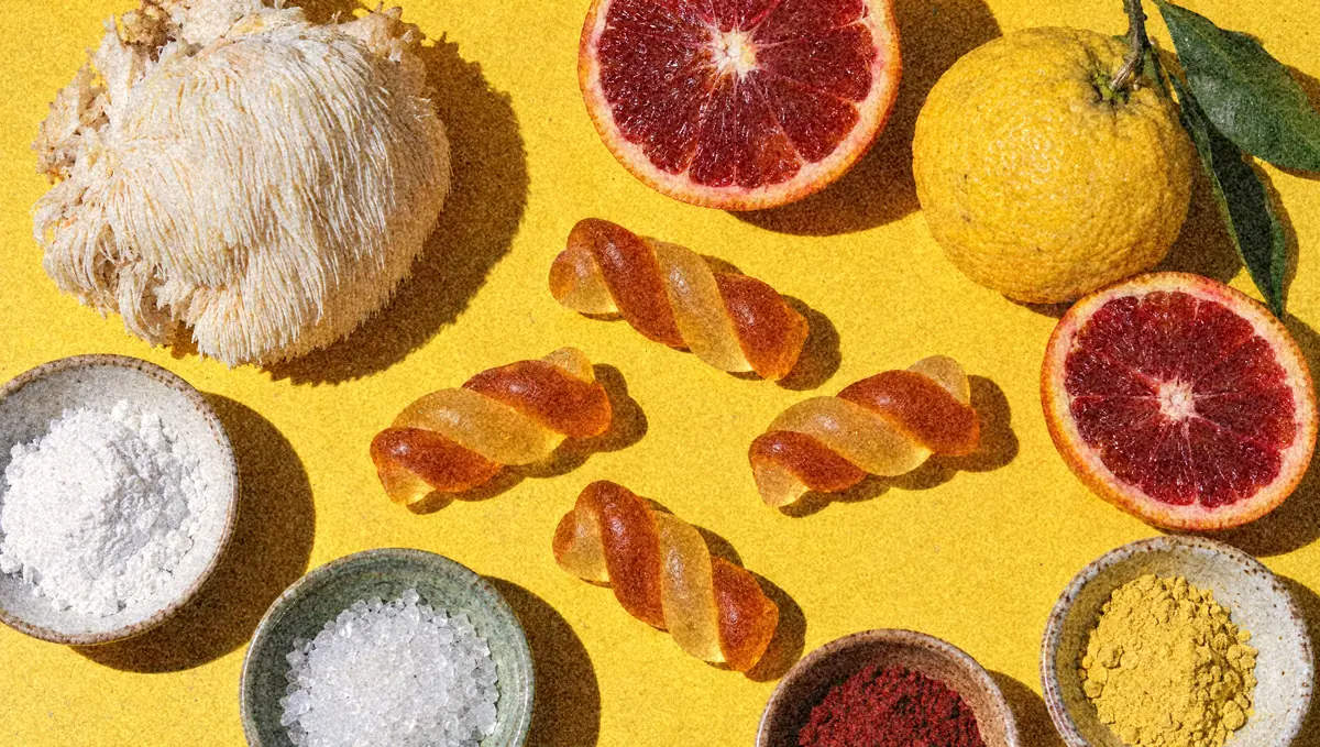 Flat lay on yellow paper: a Lion’s Mane mushroom, blood oranges, a yuzu, twist gummies and small bowls of white, red and yellow powders